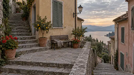 A wide stone landing between two flights of stairs holds a wooden bench with a folded cardigan and potted geraniums and lemon trees between shuttered house fronts with an unlit stair lamp on the wall in warm evening light.