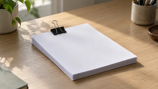 A black metal binder clip with two folding silver wire handles clamps the top edge of a neat stack of about ten blank white sheets lying flat on a plain light oak desk in soft side daylight seen from a slight high angle.