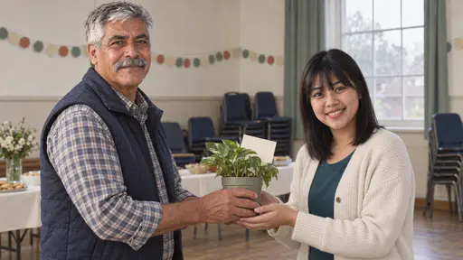 An older man in a navy gilet hands a young woman in a cream cardigan over a teal top a small potted plant with a blank cream card tucked into it as both smile with her hands cupped to receive it in warm hall light.