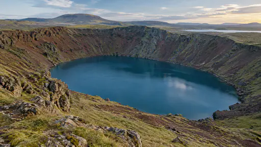 A still blue lake fills a wide volcanic crater with red-brown inner slopes and a grassy rim, seen from the rim in clear evening light.