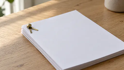 A single brass split pin paper fastener passes through a punched hole in the top-left corner of a neat stack of about ten blank white sheets lying flat on a plain light oak desk with its two flat prongs spread apart in soft side daylight.