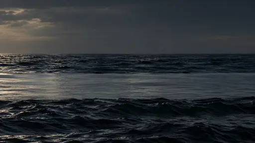 A flat calm surface slick lies in one smooth band between two rougher dark wave bands in low dark light, catching faint light.