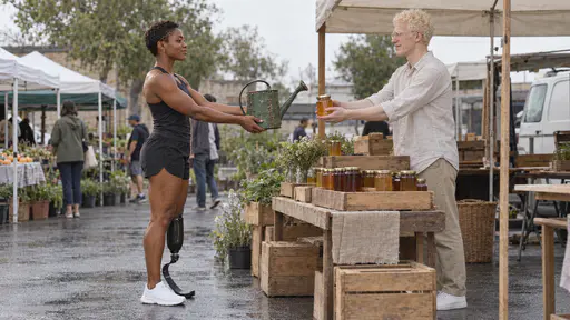 A Black woman runner in her late twenties with a carbon-fibre blade holds out a battered green enamel watering can with copper stitches at a plain market stall while a slim man with albinism in his late twenties holds out a plain jar of honey toward her in exchange, crates of jars between them in midday light.