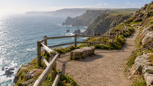 A clifftop coastal path in bright hazy light widens into a railed viewing pause with a timber rail and a single flat stone seat while the path continues clear past it along the cliff edge with a sparkling sea and pale headland beyond.