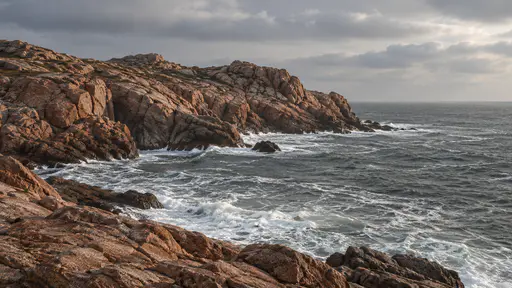 A dry rocky headland of bare red-brown stone juts into a grey sea in clear side light with white water breaking at its foot.