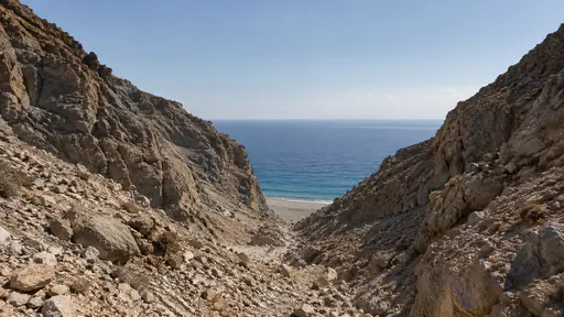 A dry stony ravine cuts between bare slopes in clear midday light and opens out onto a pale shore where it meets the sea.