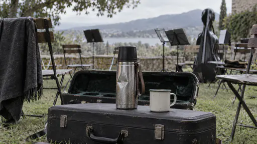 A dented steel flask stands on an instrument case with a shared mug beside it among scattered folding chairs on a lawn with music stands and a cello case blurred behind in soft shade.