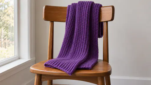 A folded violet wool scarf with a loose ribbed knit hangs over the back of a plain wooden chair beside a window against a bare wall in soft even daylight.