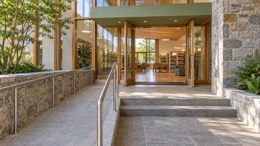 A gentle stone ramp with a continuous steel handrail climbs beside two shallow steps to a smooth threshold strip and open glass library doors in clear midday light.