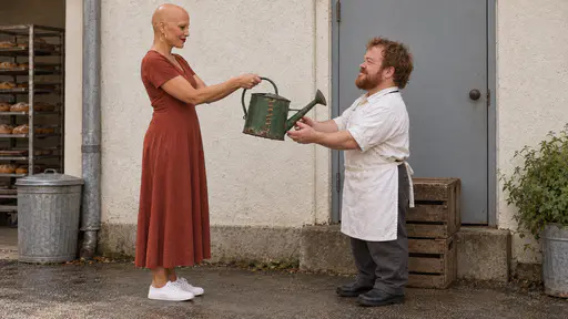 A woman with alopecia in her mid forties holds out a battered green enamel watering can with copper staple stitches as a gift at a plain bakery side door while a short-statured man in his forties with a red-brown beard reaches with both hands to take it in warm morning light.