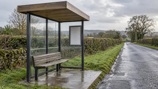 A simple bus shelter on a rural road has a plain timber roof, three glazed panels, a slatted bench and a blank timetable frame beside a wet grass verge and a single-lane road between hedgerows in soft grey light.