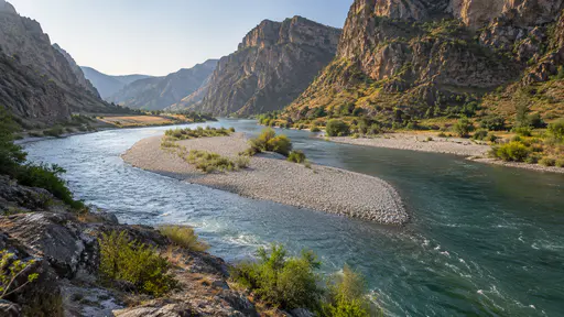 A pale gravel bar splits the canyon river into two channels that rejoin beyond it below plain rock walls in bright morning light.