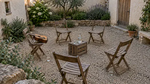 A loose circle of folding wooden chairs stands in a plaster courtyard with an acoustic guitar resting across one seat, glasses on the gravel beside two chairs and a water bottle on an upturned crate in warm early-evening light.
