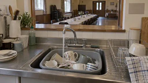 The tap over a deep sink in a community hall kitchen runs into a washing-up bowl piled with mismatched cups with a drying rack and tea towel nearby and a long table of empty chairs through the hatch in warm interior light.