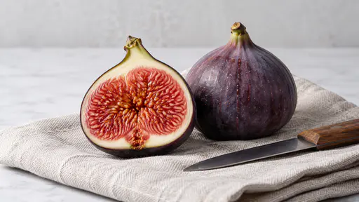 A halved ripe fig with pink-red flesh and violet skin rests beside a whole violet fig on a folded natural linen cloth with a small plain knife in bright diffuse studio light.