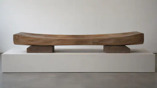A low horizontal wooden form balances on two broad flat supports on a long pale plinth against a plain wall in even gallery light.