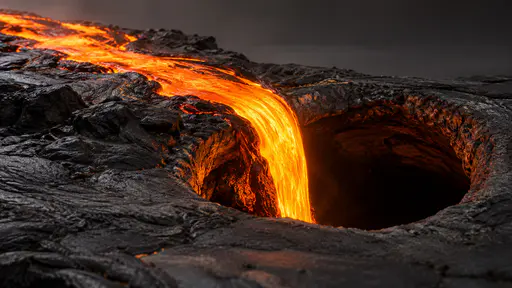 One bright molten stream pours in a smooth arc down into the dark round mouth of a lava tube in dim light.
