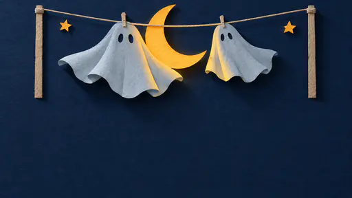 Whimsical layered paper-cut illustration of a saffron crescent moon snagged on a short washing line between two billowing ghost-shaped paper sheets, above a wide open indigo night sky — a Halloween background with copy space.