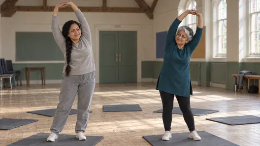 Two women, one with a long black braid in a grey tracksuit and one silver-haired in a teal tunic, stretch with raised arms on foam mats in a circle with empty mats between them in a timber-beamed hall in low morning light.