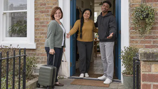 A woman with an auburn bob in a sage-green cardigan holding a small wheeled suitcase and a tote bag stands on a doorstep while a woman with a long braid in a mustard jumper and a young man in a charcoal hoodie open their arms in welcome in the blue doorway.