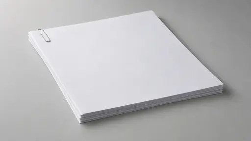 A single silver wire paperclip holds the top-left corner of a neat stack of about ten blank white sheets lying flat on a plain pale grey surface seen from a slight high angle with empty space around it.