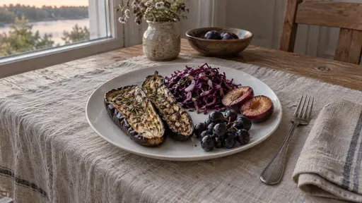 A plain white plate on a scrubbed pine table holds roasted aubergine halves with glossy violet skin, a pile of purple cabbage slaw, two plum halves and a small bunch of dark grapes with a fork and a linen napkin beside it in warm evening window light.