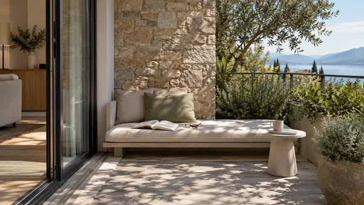 A cushioned terrace bench holds an open blank book laid face down and folded reading glasses, with a small side table and one plain cup.