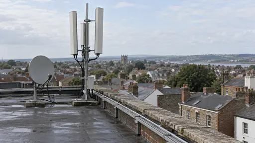 A small satellite dish and an array of three slim panel antennas on a short pole stand on a flat brick rooftop with a plain cable tray along the edge above a modest townscape of chimneys under a pale blue sky.