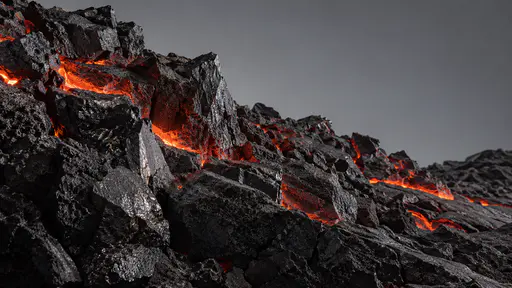 A rough fragmented lava edge of jagged black blocks tumbles down a slope in hard side light with thin glowing cracks between the blocks.