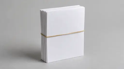 A single plain tan rubber band stretches around the middle of a neat stack of about ten blank white sheets standing on its long edge on a plain pale grey surface seen from a slight high angle with empty space around it.