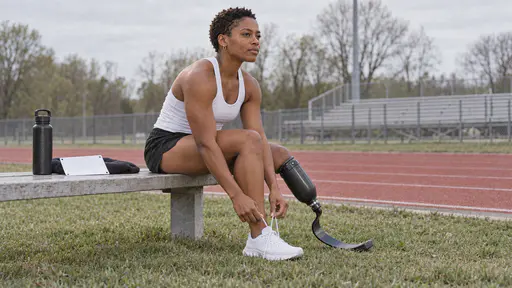 A lean woman with close-cropped hair and silver hoops and a black carbon-fibre running blade on her right leg sits on a low bench beside the track lacing her left running shoe with both hands, a water bottle and a blank bib beside her in soft morning light.