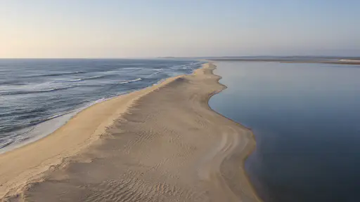 A long narrow sand spit runs between open sea on one side and a still flat lagoon on the other toward a low horizon in even morning light.
