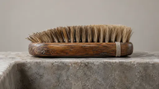 A wooden-handled scrubbing brush rests bristles-up on a stone sink edge with its bristles worn to a slant, its wooden back darkened and smoothed and a replaced cord binding on the handle in soft diffuse light.