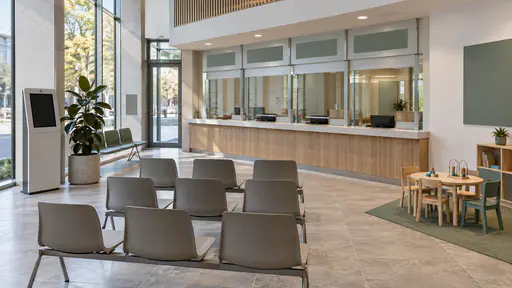 A service-desk waiting area has plain moulded chairs facing a long counter with several closed windows, a ticket machine with a blank screen, a rubber plant and a children's play corner with a small table in even office light.