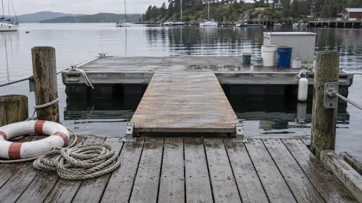 A flat wooden platform spans the gap between a small jetty and a floating pontoon with a coil of rope and a lifebuoy on the jetty and paint pots on the pontoon over calm water in soft overcast light.