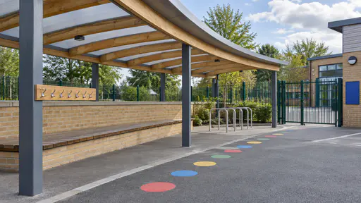 A sheltered school pickup point has a long curved canopy over a low brick wall used as a bench, a post with small coat hooks, a bike rack and painted coloured circles on the tarmac with the school gate behind in bright afternoon light.