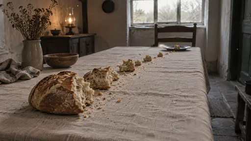 A single round loaf is broken into pieces laid along a long plain table with the pieces getting smaller toward the far end and the smallest piece on a tin plate beside an empty chair at the last place in warm lamp light.