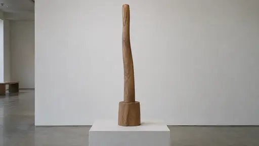 One slender tall wooden form rises from a short solid wooden base on a pale plinth against a plain wall in even gallery light.