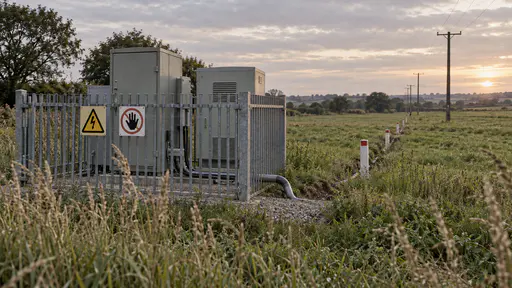 A small fenced enclosure of grey metal cabinets stands in the corner of a field behind a low steel fence with pictogram-only warning signs while cable markers lead across the grass toward a line of poles in golden hour light.