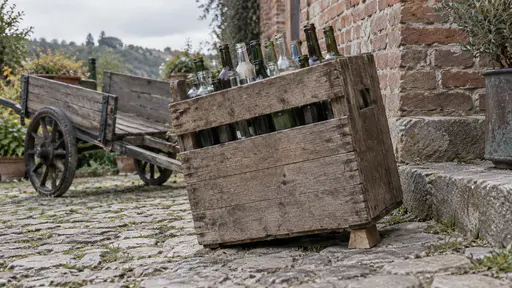 A tall wooden crate of bottles is steadied by a small plain wooden wedge pushed under its low corner on a sloping yard with a hand cart and a brick wall behind in bright afternoon light.
