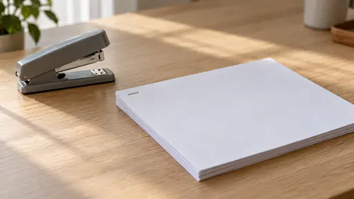 A single small silver staple is driven through the top-left corner of a neat stack of about ten blank white sheets on a plain light oak desk beside an open plain grey desktop stapler in soft side daylight.