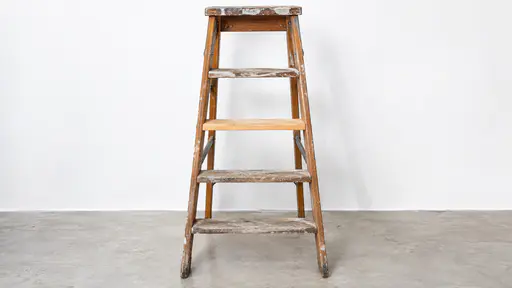 A wooden step ladder stands open against a plain white wall with rungs worn pale in the middle, one fresh bright rung of newer wood replacing a lost one and a stain of old paint on the top step in bright afternoon light.