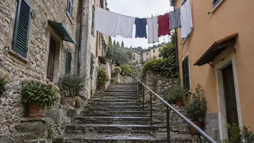 A narrow stepped alley with a metal handrail runs between two buildings with an overhead laundry line of white sheets and coloured shirts strung between two upper windows above damp stone steps in bright overcast light.