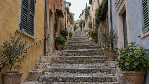 A stepped cobbled lane climbs between tall shuttered house fronts in ochre, rust and pale blue plaster with a worn iron handrail and potted plants on the steps rising toward a bright sky in early morning side light.