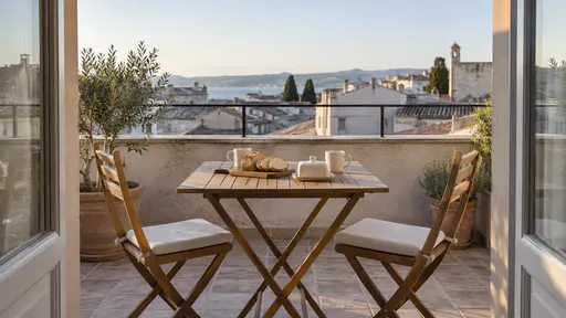 A small tiled terrace holds a table set with a bread board, covered butter dish, two plain cups and two chairs beside a low railing.