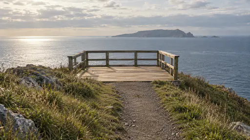 An empty timber lookout platform with a low railing stands at the top of a grassy headland overlooking a calm sea and a distant island in clear late-afternoon light.