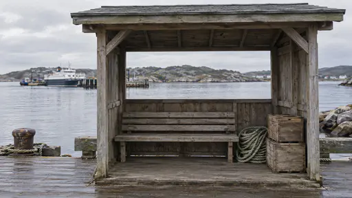 A small open-fronted timber waiting shelter with a plain bench facing calm harbour water stands at a harbour edge with coiled rope and crates beside it in soft morning light.