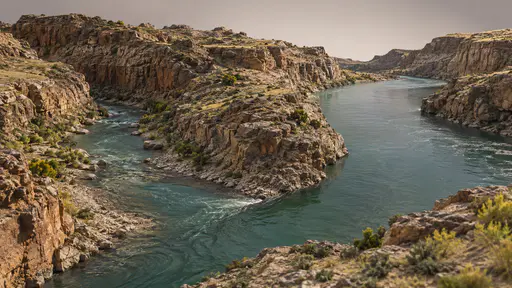 A smaller tributary stream joins the main canyon river at a clear angled confluence between rock banks in even afternoon light.