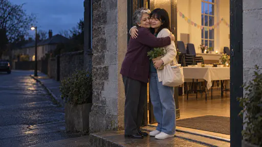 A young woman in a cream cardigan over a teal top holding a cloth bag and a small potted plant hugs an older woman in a plum cardigan in a lit hall doorway at blue dusk with warm light spilling onto the step.