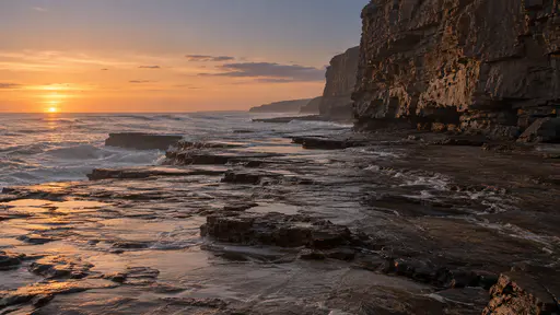 A flat wave-cut rock platform stretches at the sea edge beneath a high bare rock face in low evening light, thin water sheeting across its surface.